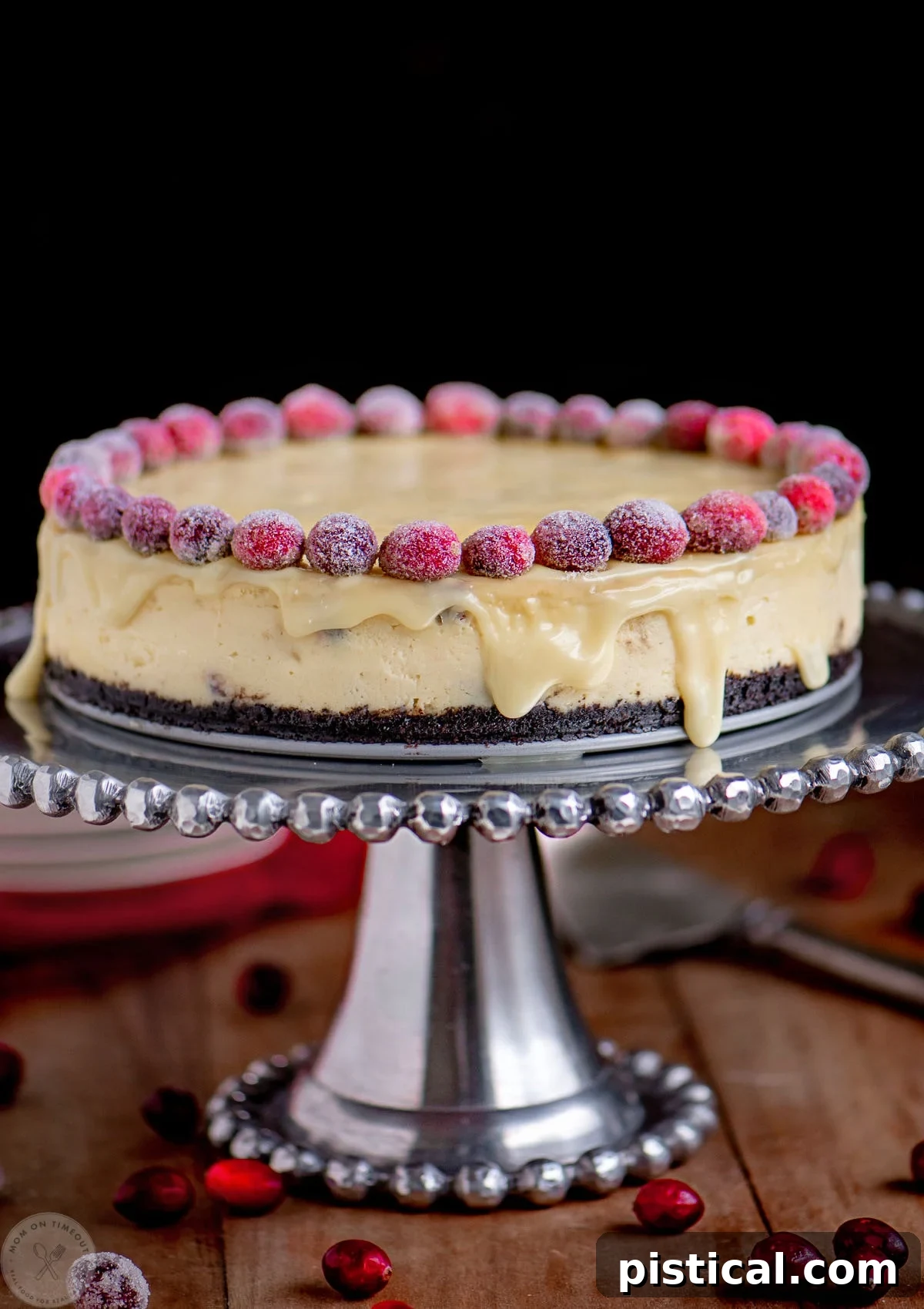 A stunning White Chocolate Cranberry Cheesecake presented on a silver cake stand, adorned with glistening sugared cranberries and topped with a luxurious white chocolate ganache, ready for holiday festivities.
