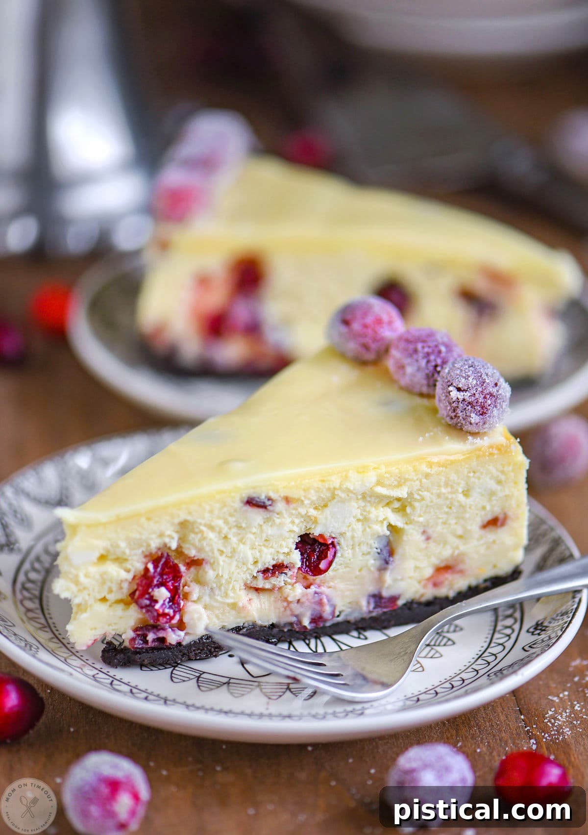 A perfectly sliced piece of Cranberry White Chocolate Cheesecake on a round white plate, garnished with sugared cranberries and a drizzle of white chocolate ganache, showcasing its rich layers and festive appeal.