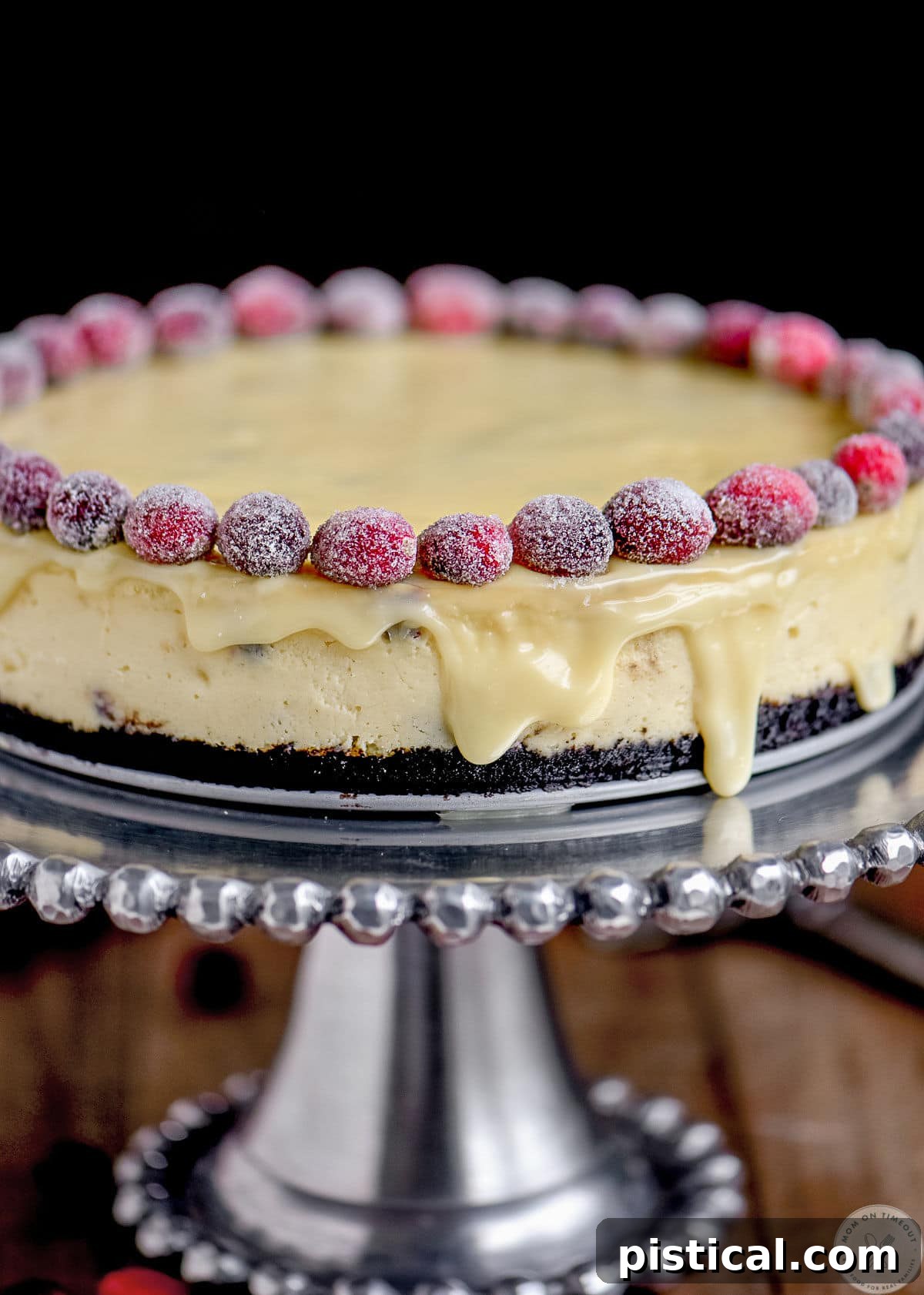 A close-up shot of the decadent White Chocolate Cranberry Cheesecake, richly textured with cranberries and a smooth ganache, sitting on a silver cake stand.