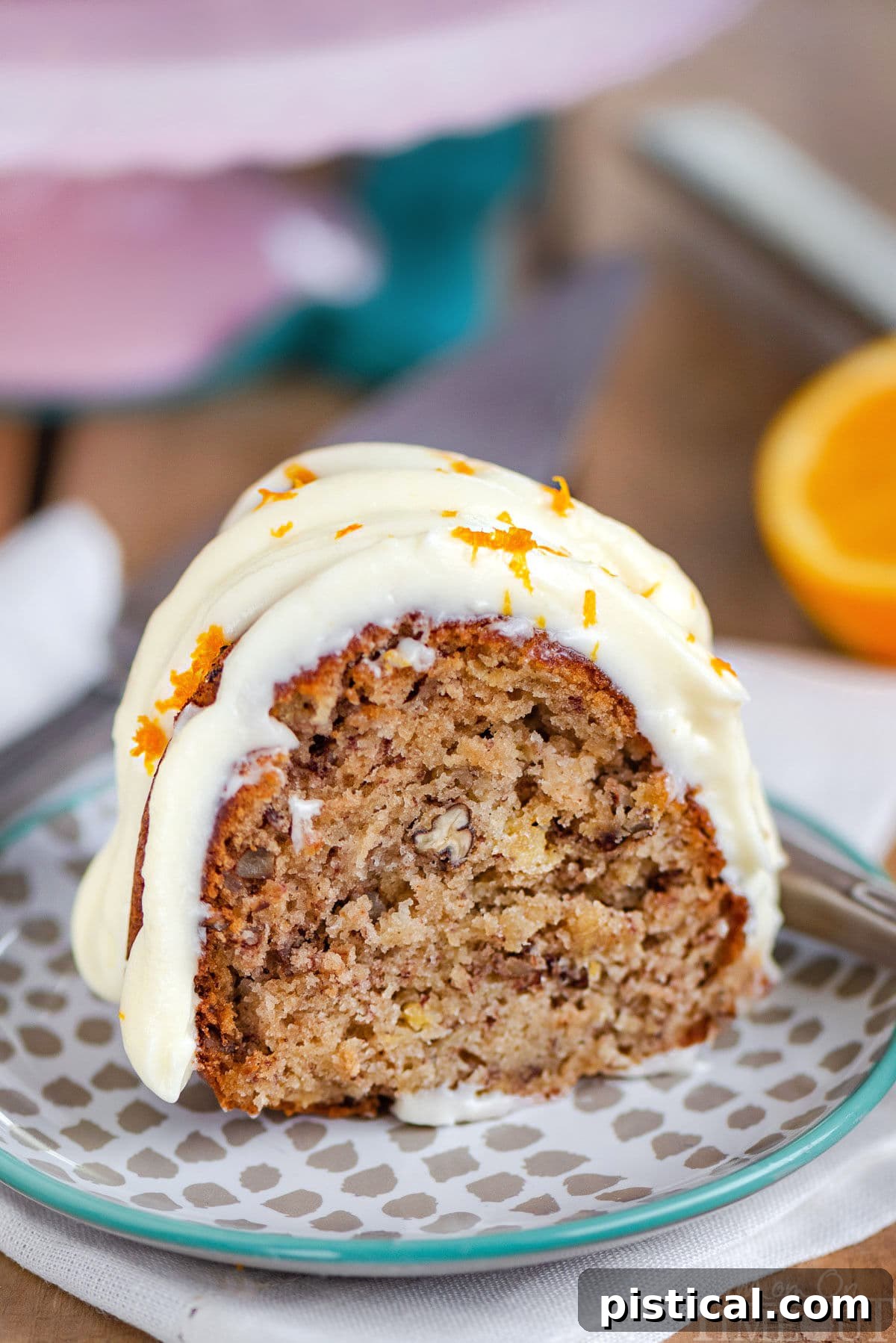 A beautifully presented slice of Hummingbird Bundt Cake, generously topped with a creamy, drizzled cream cheese frosting, resting on a small dessert plate in front of a charming pink cake stand.