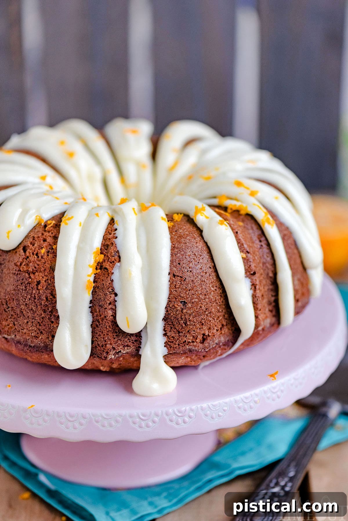 A stunning full Hummingbird Bundt Cake displayed on a pink cake stand, showcasing the elegant cream cheese frosting artfully drizzled over its intricate ridges and topped with fresh orange zest.