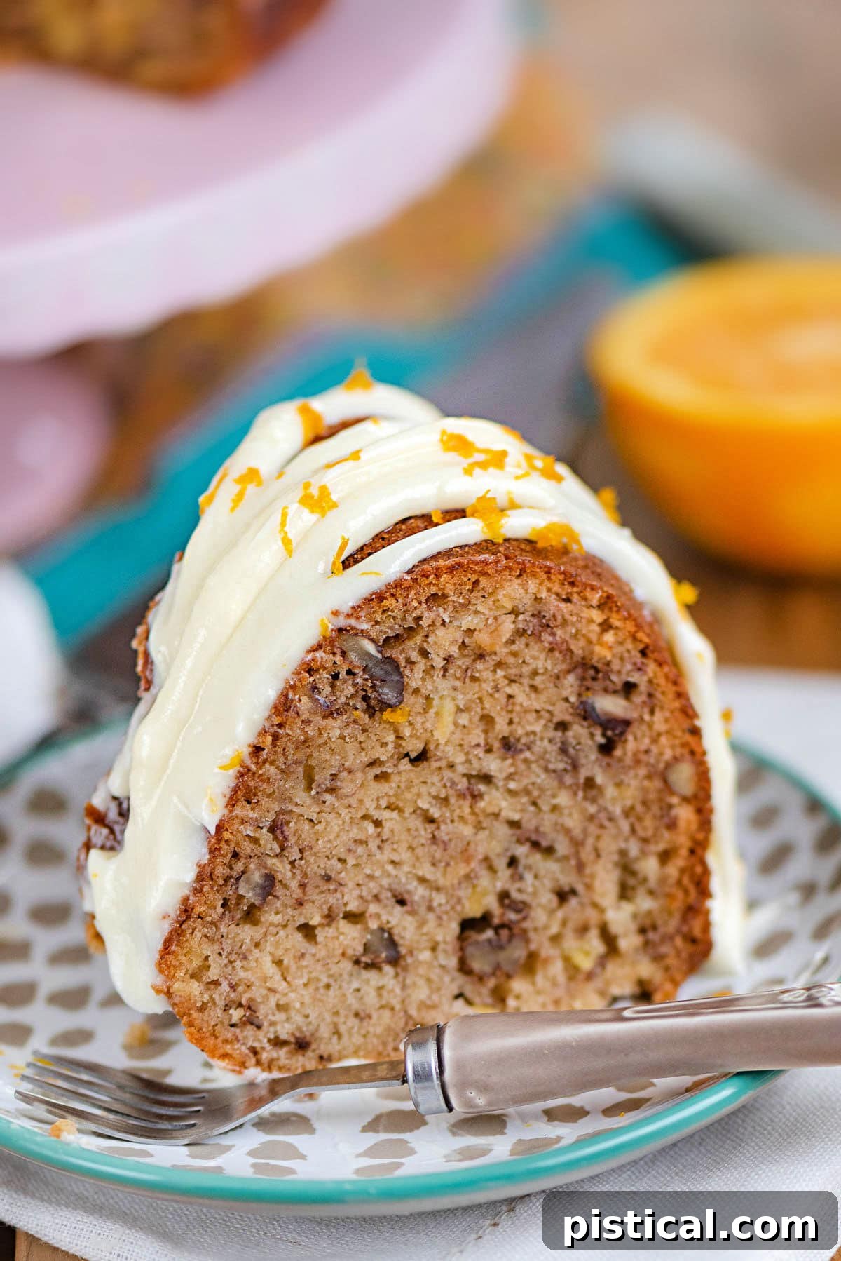 A perfectly baked slice of Hummingbird Bundt Cake on a small dessert plate, beautifully topped with a creamy, drizzled cream cheese frosting, elegantly placed in front of a charming pink cake stand.
