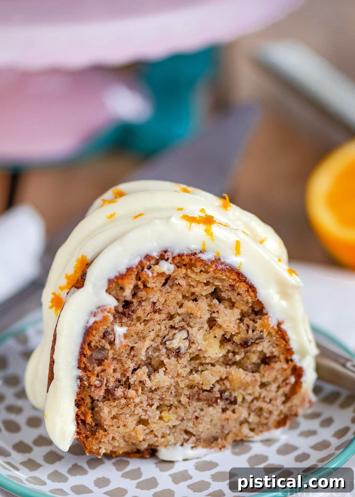 A close-up of a delectable slice of Hummingbird Bundt Cake, richly coated with creamy cream cheese frosting and garnished with orange zest, presented on a small dessert plate in front of a lovely pink cake stand.