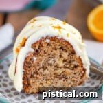 A charming image of a slice of Hummingbird Bundt Cake on a small dessert plate, placed in front of a pink cake stand. The cake is beautifully topped with a cream cheese frosting.
