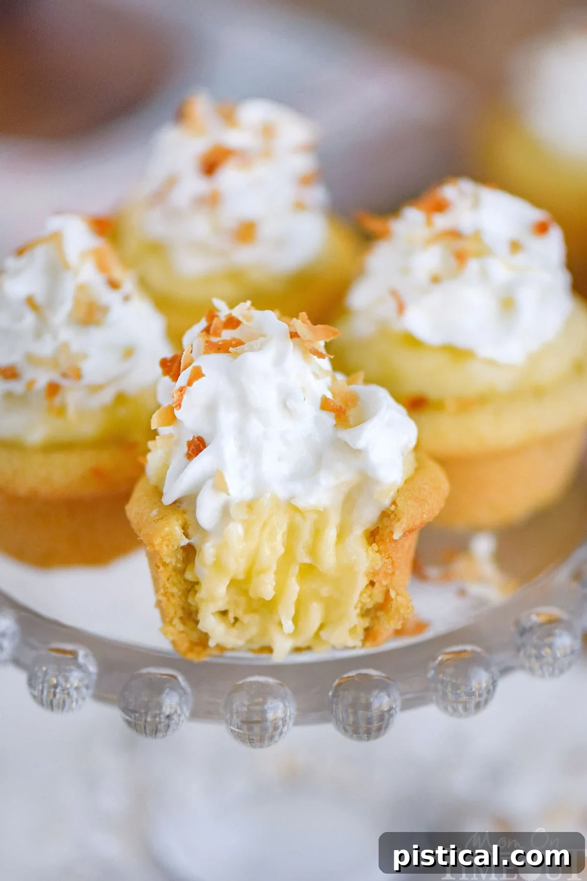 Four mini coconut cream pie cookie cups, meticulously crafted and arranged on an elegant glass cake stand. Each cup is generously topped with a swirl of fresh whipped cream and a sprinkle of golden toasted coconut flakes. The front cookie cup reveals a delightful cross-section, showing the creamy coconut custard filling within its crisp sugar cookie shell, inviting a delicious bite.