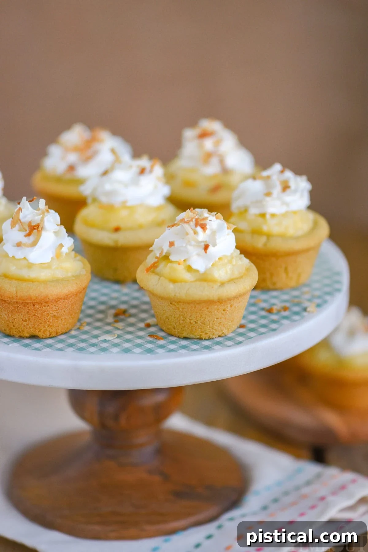 Six perfectly arranged mini coconut cream pie cookie cups presented on an elegant marble cake stand with a sturdy wooden base. Each dessert features a golden sugar cookie base filled with creamy coconut custard, topped generously with whipped cream and lightly toasted coconut flakes, ready to be enjoyed.