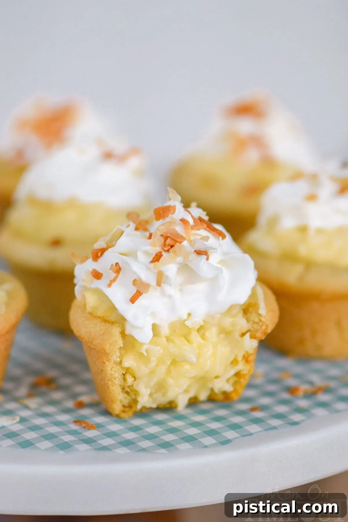 A close-up view of a coconut cream pie cookie cup, artfully cut in half to reveal its layers. The crisp sugar cookie crust cradles a luscious, creamy coconut custard filling. It's elegantly topped with a generous swirl of whipped cream and a scattering of golden-brown toasted coconut flakes. The cookie cup sits on a white marble cake stand, emphasizing its perfect texture and inviting appeal.