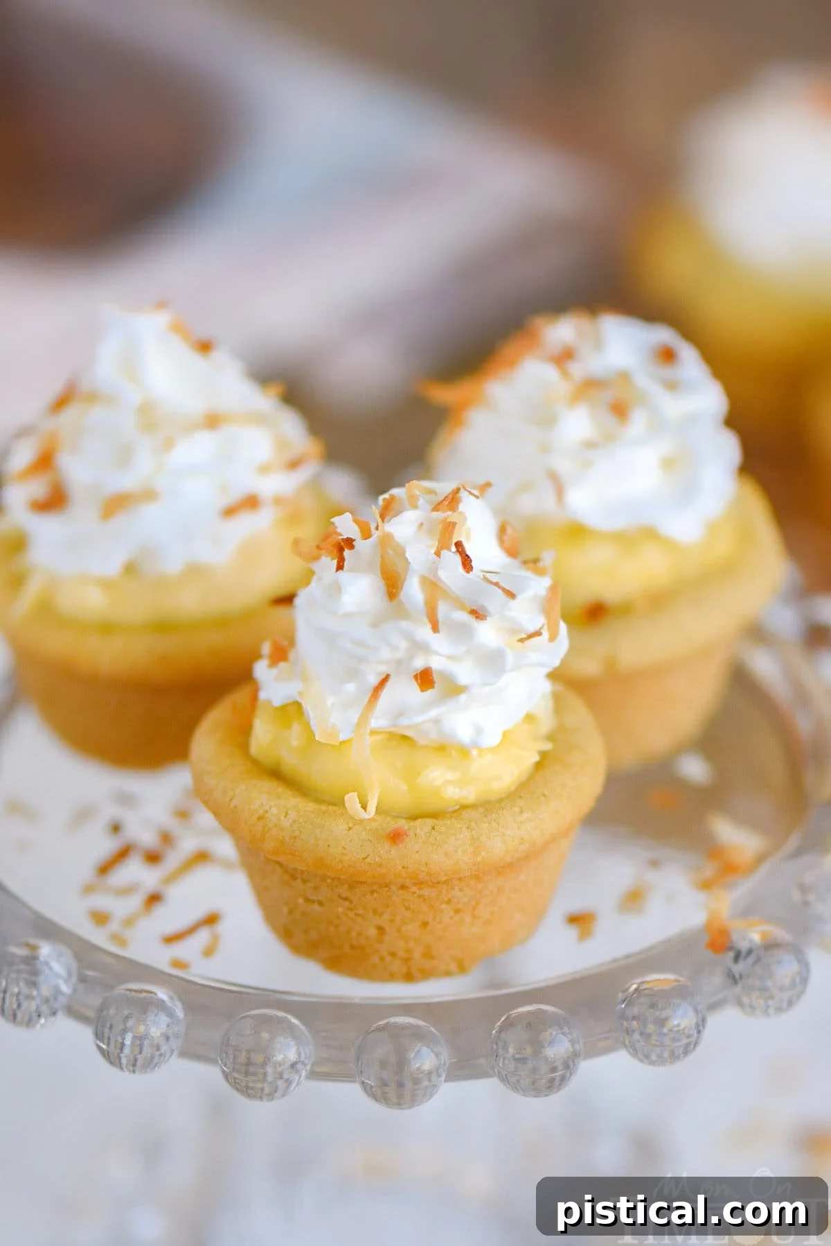Three beautifully presented coconut cream pie cookies cups displayed on a glass cake stand. Each individual dessert is generously topped with a swirl of airy whipped cream and a sprinkle of golden toasted coconut, showcasing their appealing presentation and irresistible texture.
