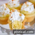 Four mini coconut cream pies on glass cake stand topped with whipped cream and toasted coconut. The front one has a bite taken out of it.