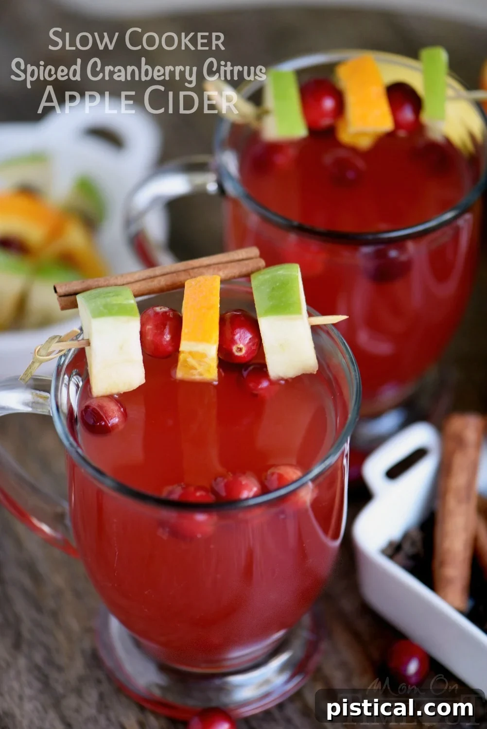 Two glass mugs filled with steaming Slow Cooker Spiced Cranberry Citrus Apple Cider, garnished with fresh apple slices, cranberries, and orange zest, resting on a rustic wooden board, emphasizing a cozy autumnal scene.