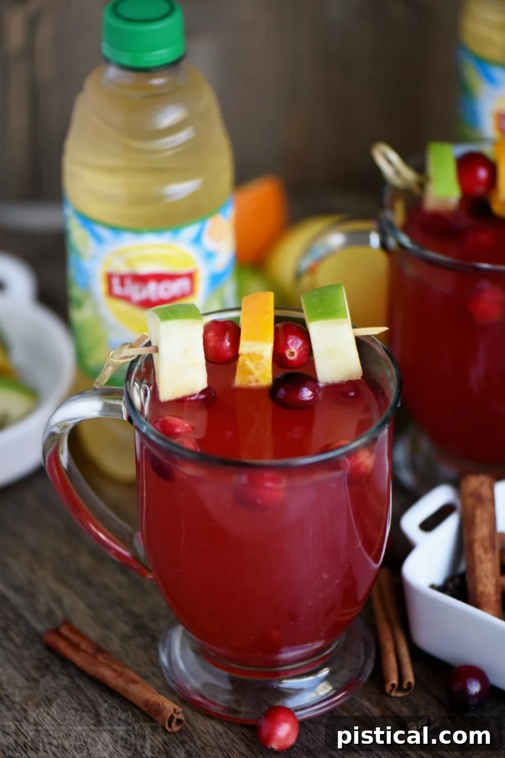 A close-up photograph of a clear glass mug filled with amber-hued spiced cranberry apple citrus cider, beautifully adorned with a single cinnamon stick. The mug is surrounded by a scattering of fresh, ruby-red cranberries, evoking a sense of warmth, comfort, and festive seasonal charm.