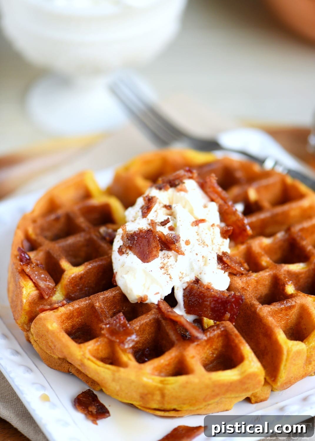 A close-up of a Pumpkin Spice Maple Bacon Waffle topped generously with whipped cream, chopped maple bacon, and a sprinkle of pumpkin pie spice.