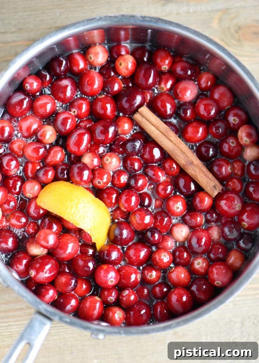 Close-up of homemade cranberry sauce simmering with orange slices and cinnamon stick, showing cranberries bursting.