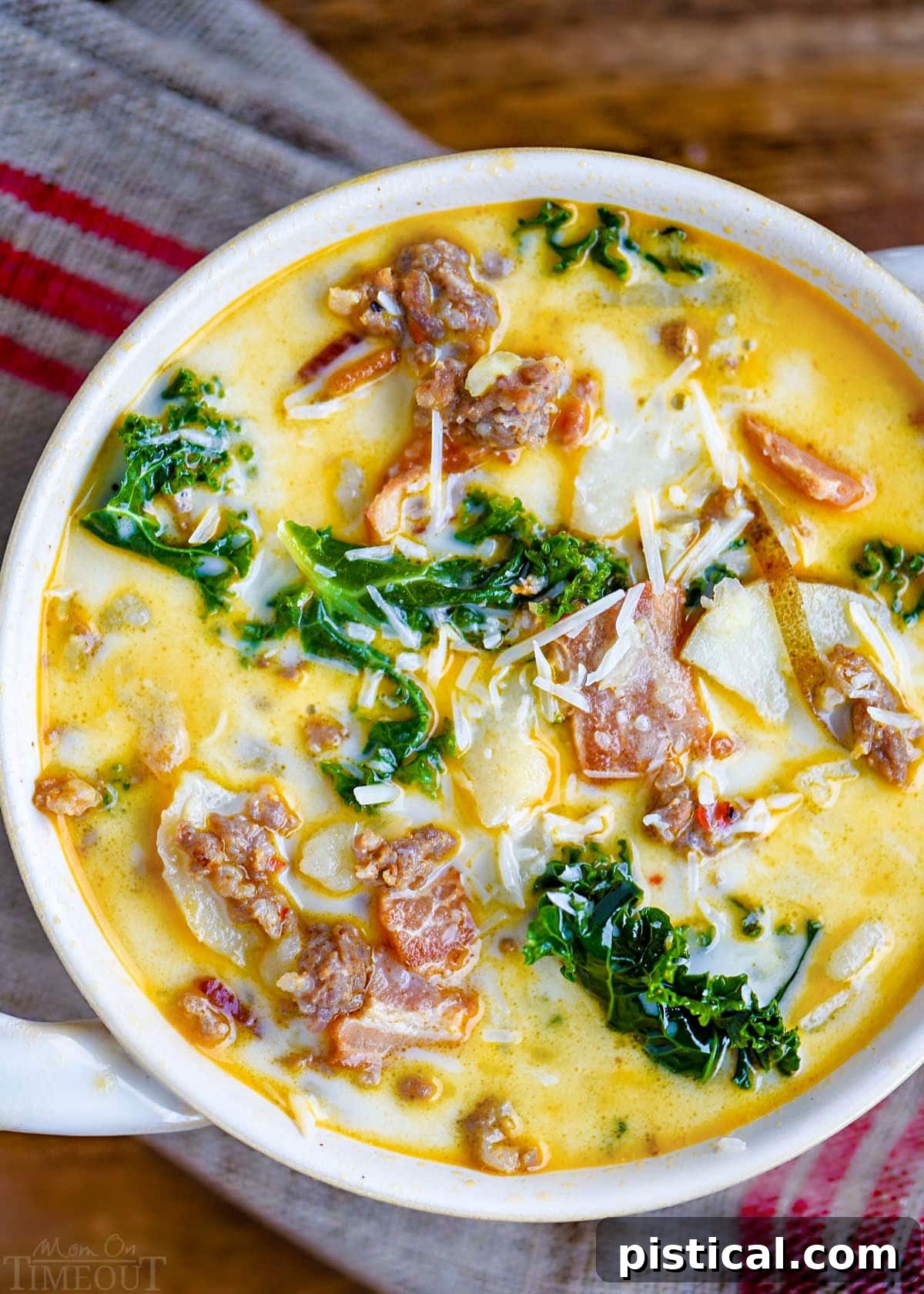 top down view of a bowl of zuppa toscana soup in a white bowl with handles. Soup is topped with Parmesan cheese and is sitting on a brown and red linen napkin.