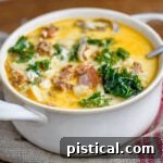 white bowl with handles filled with zuppa toscana soup and topped with cheese. Kale, sausage and bacon can be seen in the soup.
