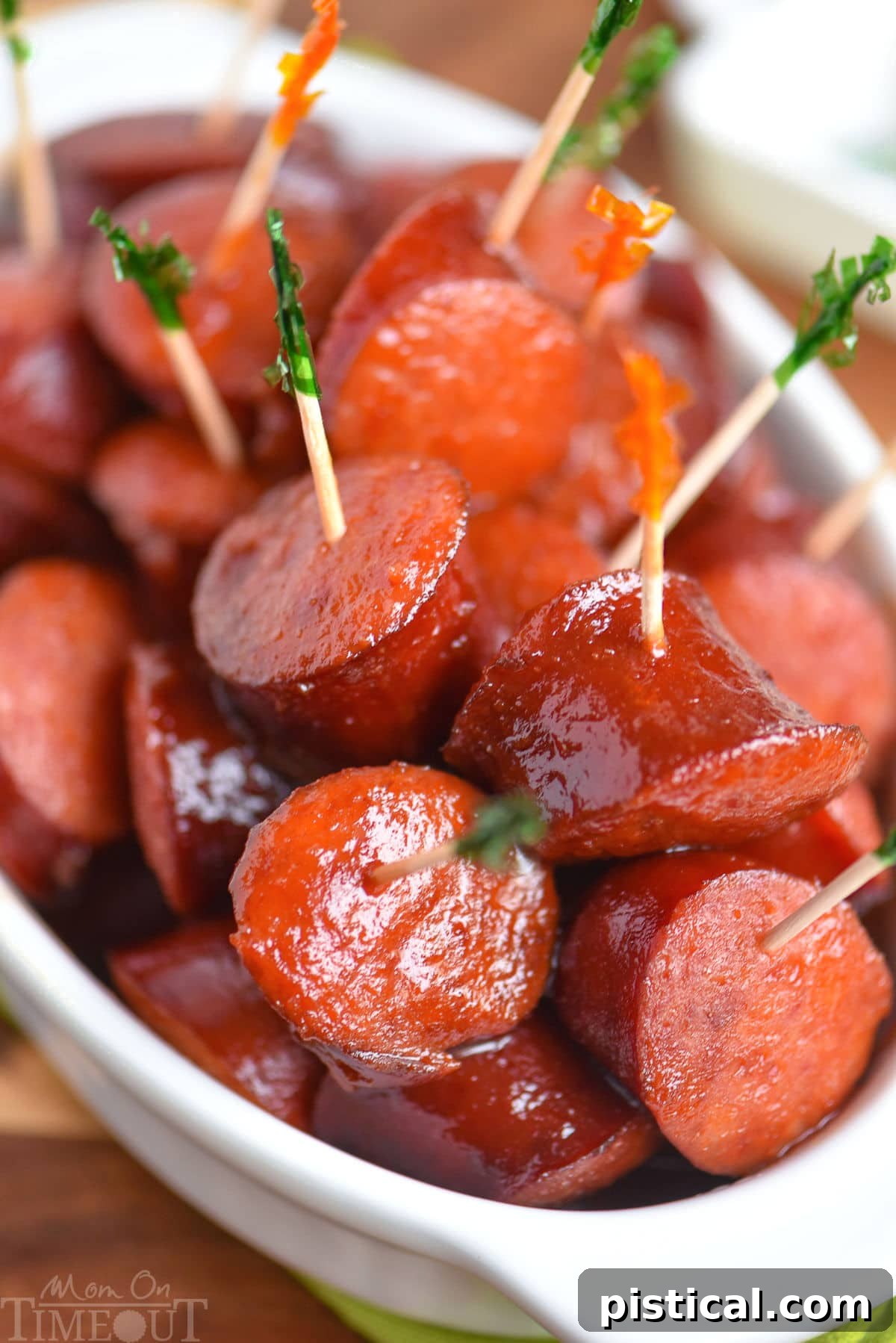 Pieces of kielbasa have been cooked in a slow cooker in a mouthwatering sauce and are shown in a white bowl with toothpicks in them.