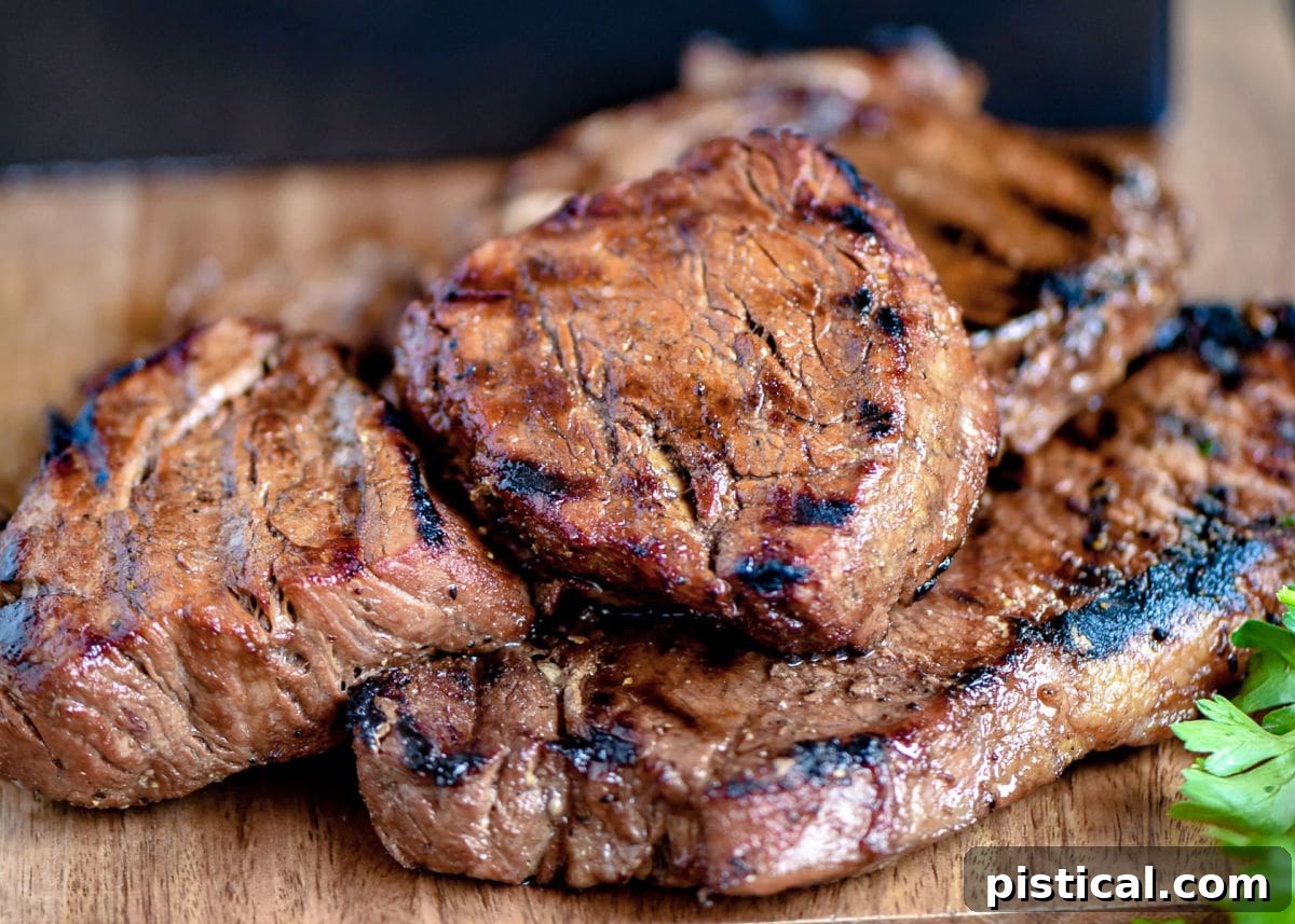 Cooked steaks with beautiful grill marks on a wooden cutting board, ready to be sliced and served, with fresh herbs in the background.