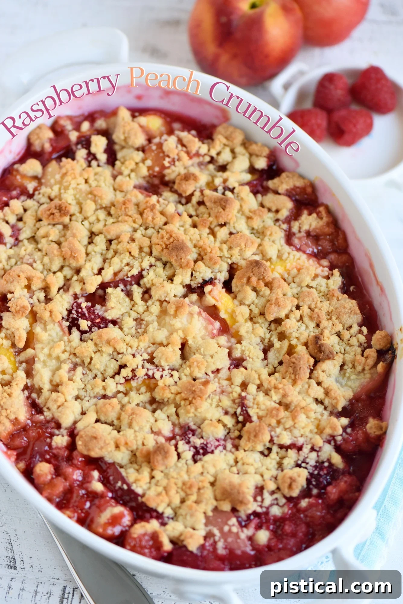 A freshly baked Raspberry Peach Crumble in a decorative baking dish, garnished with whole fresh peaches and raspberries