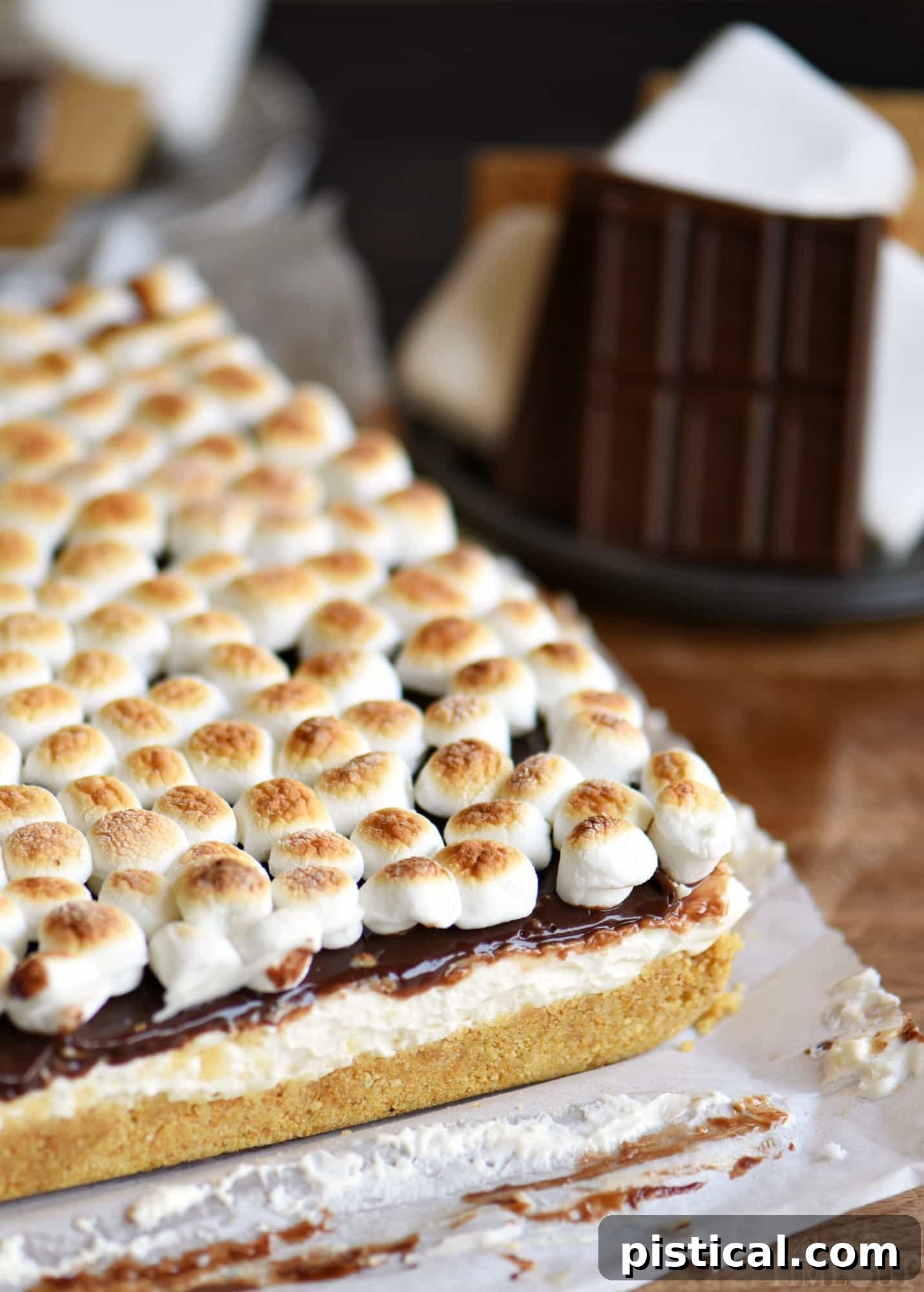 No-bake s'mores cheesecake bars in a baking pan, ready to be sliced, showing the distinct layers of crust, cheesecake, and ganache.