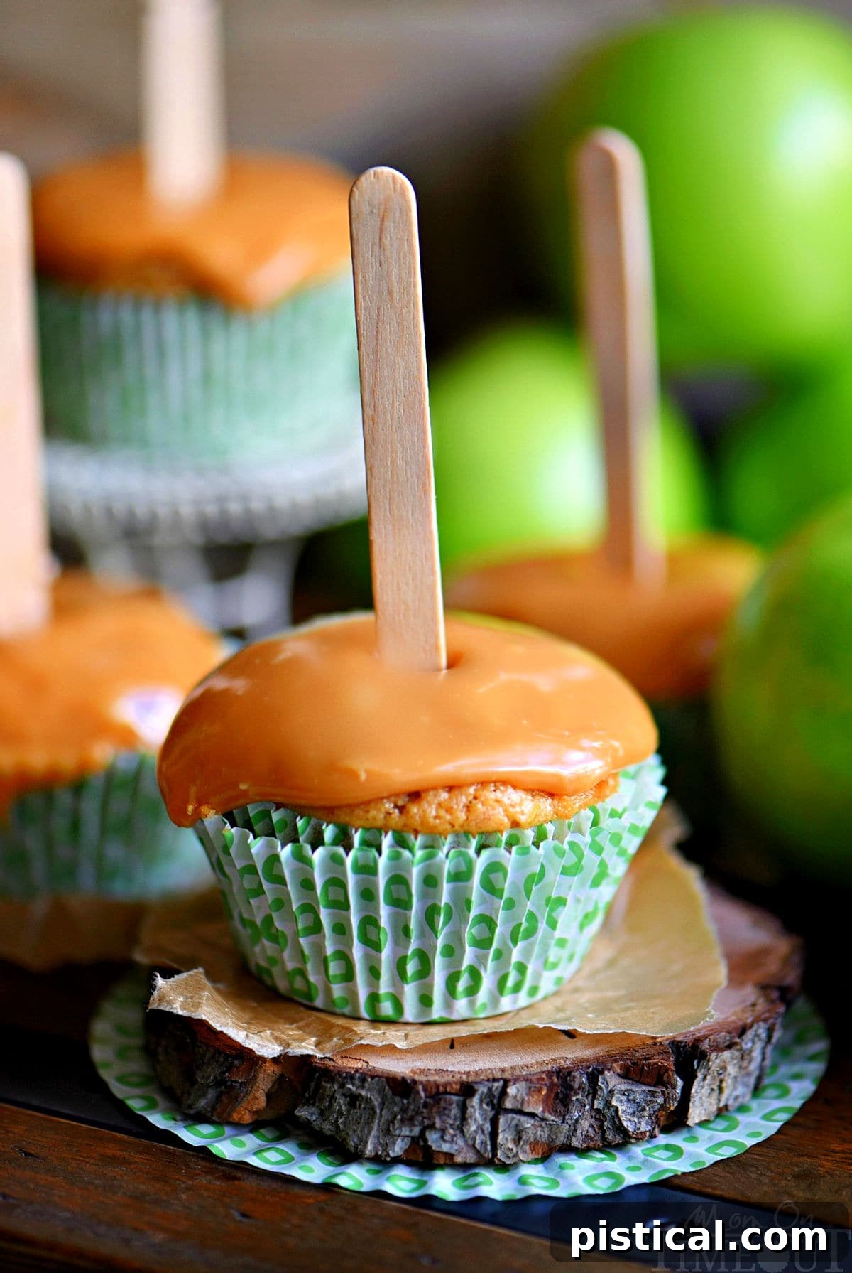 A delicious apple cupcake topped generously with a swirl of rich caramel frosting and a whimsical popsicle stick. Several other equally tempting cupcakes are blurred in the background, creating an inviting autumn scene.