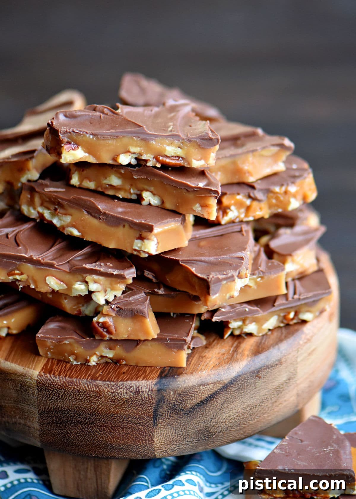 a large stack of toffee made with pecans and milk chocolate pile high on a dark wood round board.