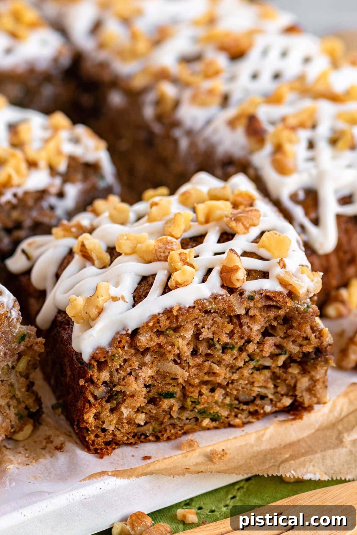 A delectable slice of zucchini spice cake, generously topped with creamy white icing and a sprinkle of walnuts, resting on a white board with the remaining cake blurred in the background.