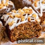 piece of zucchini spice cake on white board with the rest of the cut up snack cake behind it. The cake is topped with a cream cheese icing and walnuts.