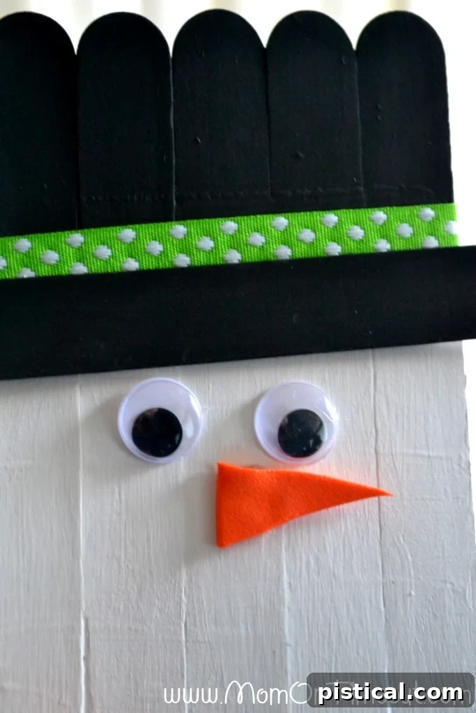 Step-by-step guide: Adding googly eyes, an orange foam nose, and a Sharpie mouth to the craft stick snowman face.