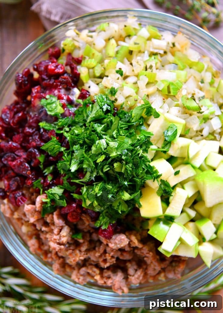 Bowl showcasing fresh ingredients for sausage stuffing: bread cubes, herbs, apples, cranberries, and raw sausage.