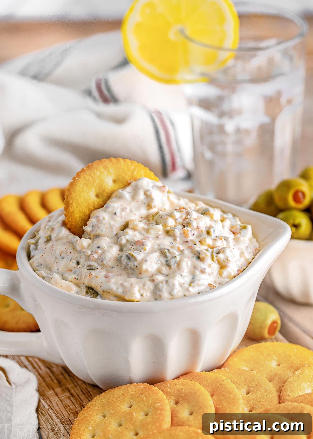 Creamy olive dip in a small white bowl on a wooden board, surrounded by an assortment of crackers and a side dish of green olives.
