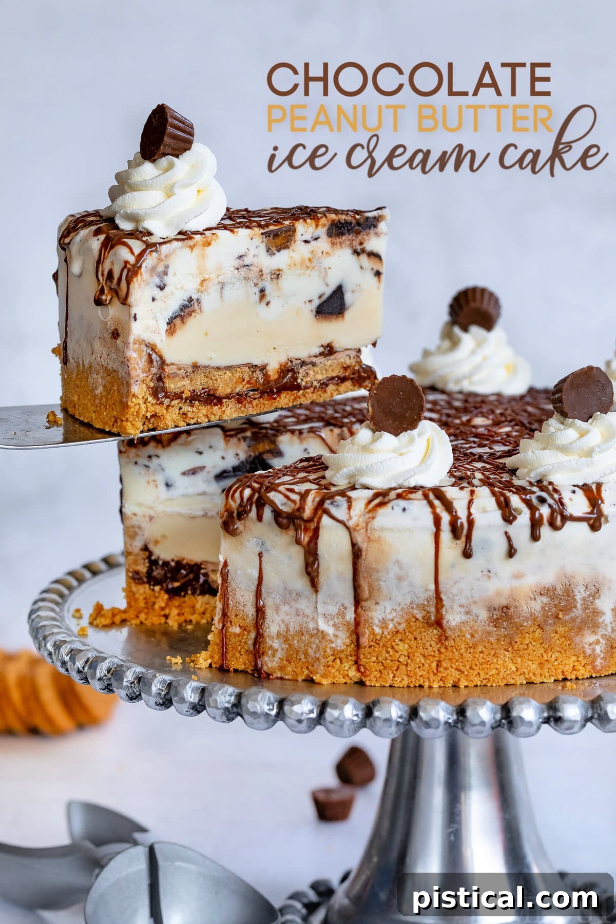 A slice of Chocolate Peanut Butter Ice Cream Cake being removed from a larger cake on a metal stand, showcasing the distinct layers of crust, fudge, Reese's, and two types of ice cream, topped with whipped cream and mini Reese's.