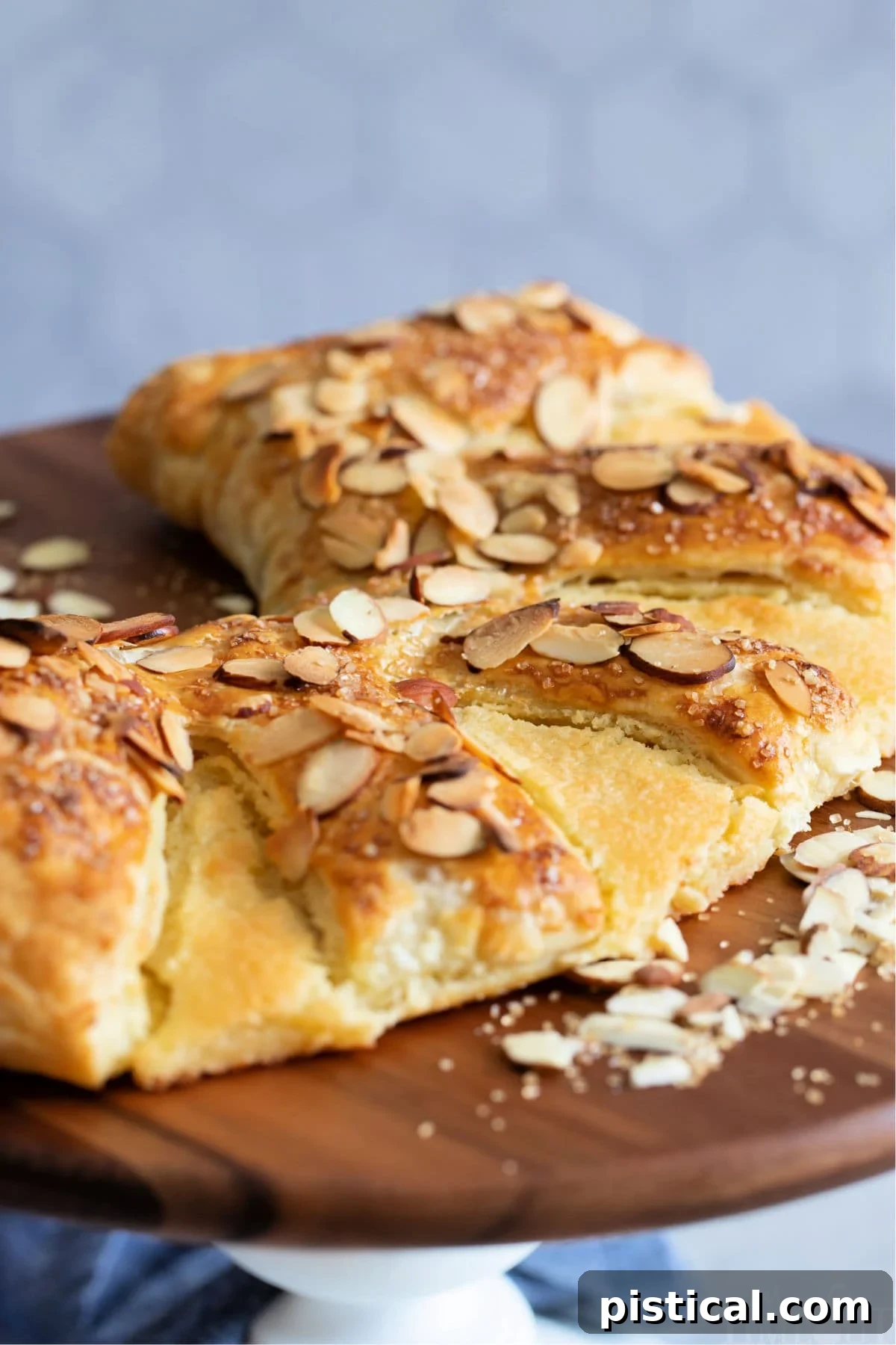 A beautifully crafted Almond Bear Claw pastry presented on a white cake stand with a natural wood top, highlighting its golden crust and delicious almond topping.