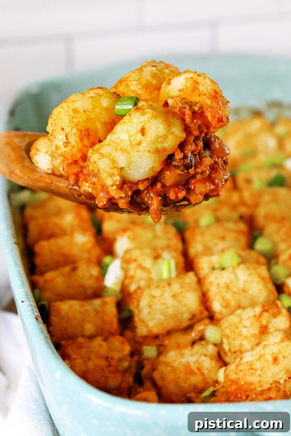 A large wooden spoon scooping out a hearty portion of Taco Tater Tot Casserole from a white baking dish, showcasing the layers of crispy tater tots, melted cheese, and the savory taco meat filling. The casserole is steaming hot and inviting.