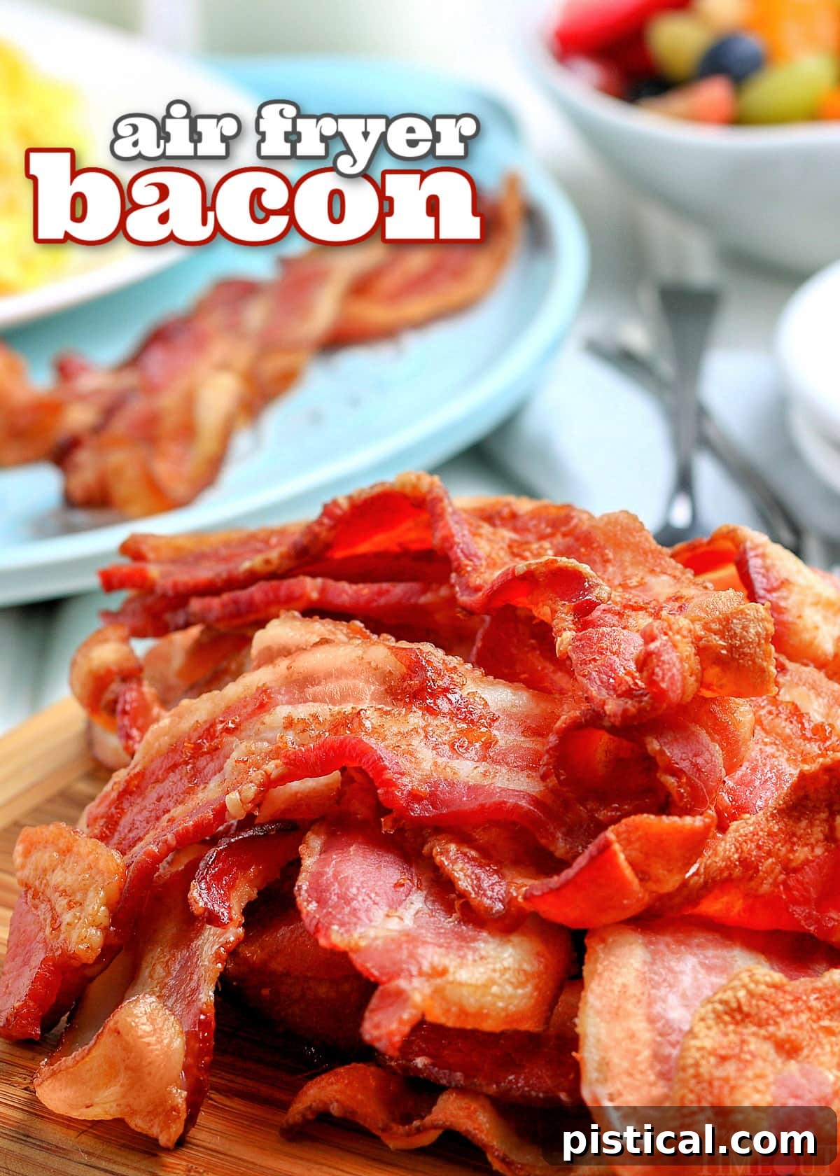 Piled high crispy air fryer bacon on a wooden board with a blue plate of eggs and bacon and a bowl of fruit in the background.