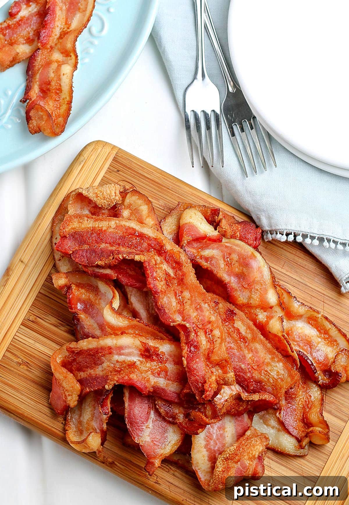 Top-down view of perfectly cooked air fryer bacon piled high on a wooden cutting board, with a stack of white plates to the side.