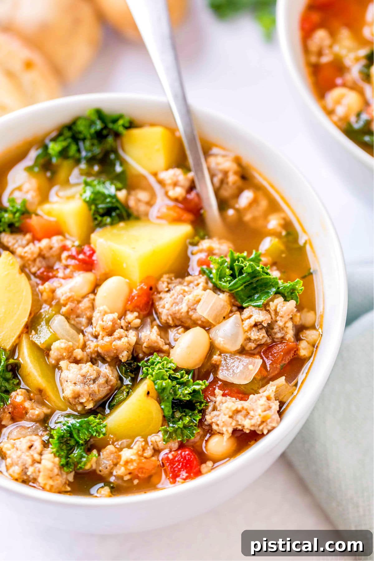A close-up view of the rich and inviting Sausage Kale Soup, showcasing the ingredients like white beans, tender potatoes, and vibrant kale, all nestled in a white bowl with a spoon ready for tasting.