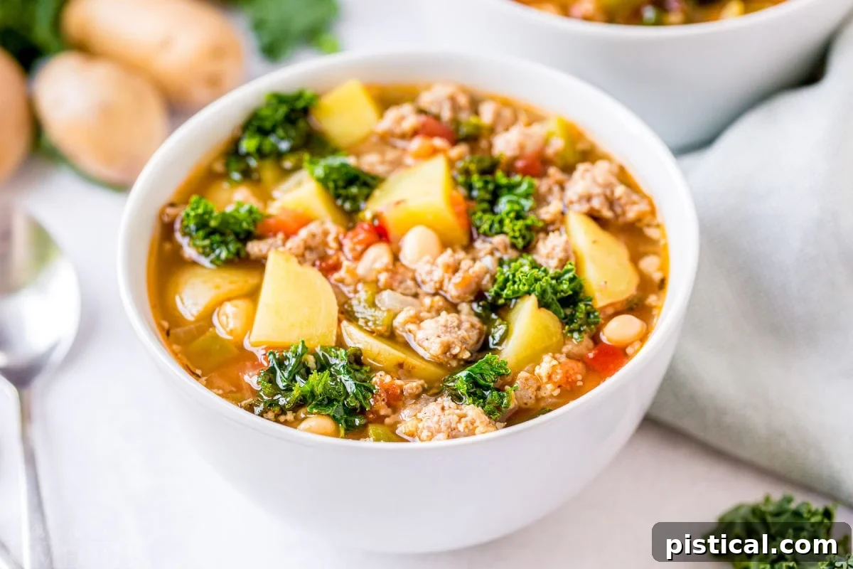 A comforting bowl of Sausage Kale Soup served on a white background, with a spoon resting beside it, and blurred potatoes and another bowl in the soft focus background.
