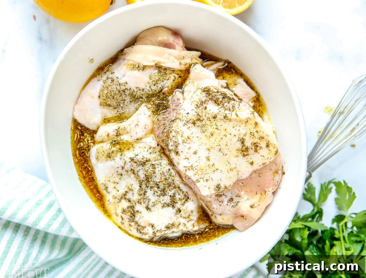 Greek chicken recipe with marinade poured over chicken thighs in a white bowl, accompanied by a green and white striped towel, lemons, and parsley for a fresh, authentic presentation.