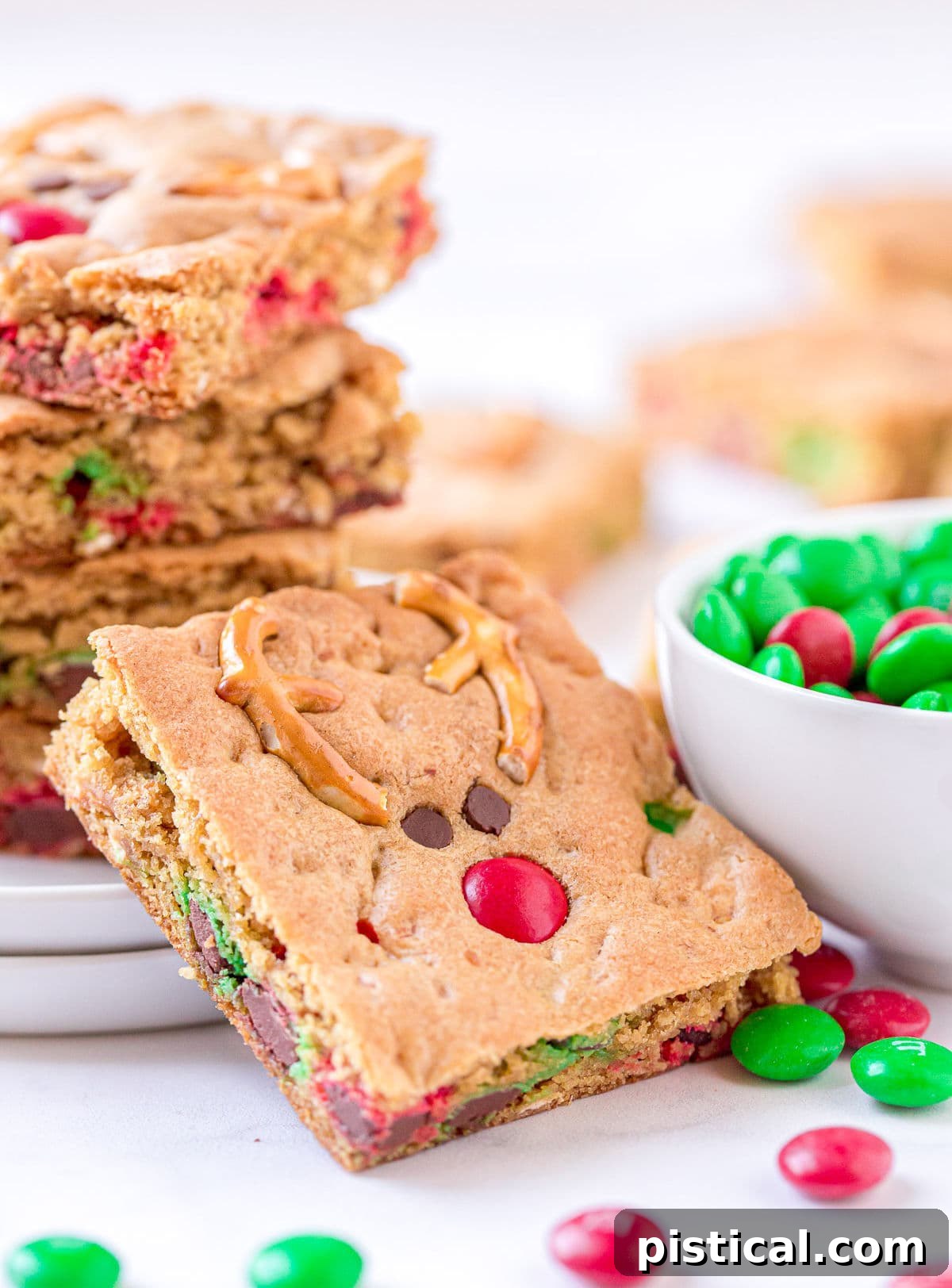 A single Reindeer Cookie Bar leans against a white plate, which holds more stacked cookie bars, next to a white bowl filled with red and green M&M's.