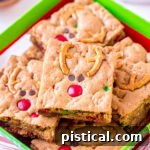 Reindeer cookie bars decorated with pretzels, M&M's, and mini chocolate chips sitting in a Christmas box.