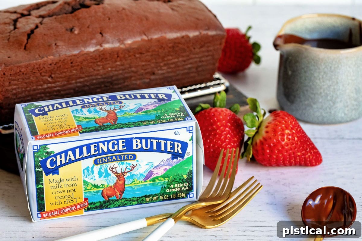 Decadent Chocolate Loaf 6 A sumptuous chocolate pound cake, perfectly sliced, with a box of Challenge Butter prominently displayed in front. Accompanying the scene are a couple of elegant forks and a scattering of fresh strawberries, highlighting the quality ingredients used.