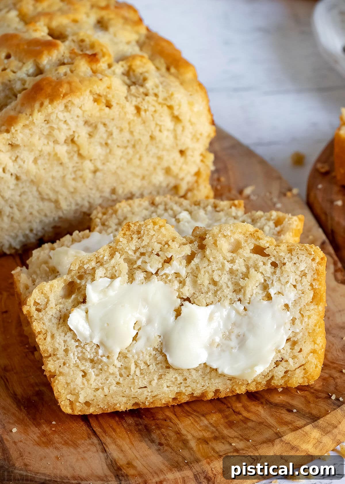 A slice of beer bread with melted butter, leaning against a whole loaf on a wooden cutting board.