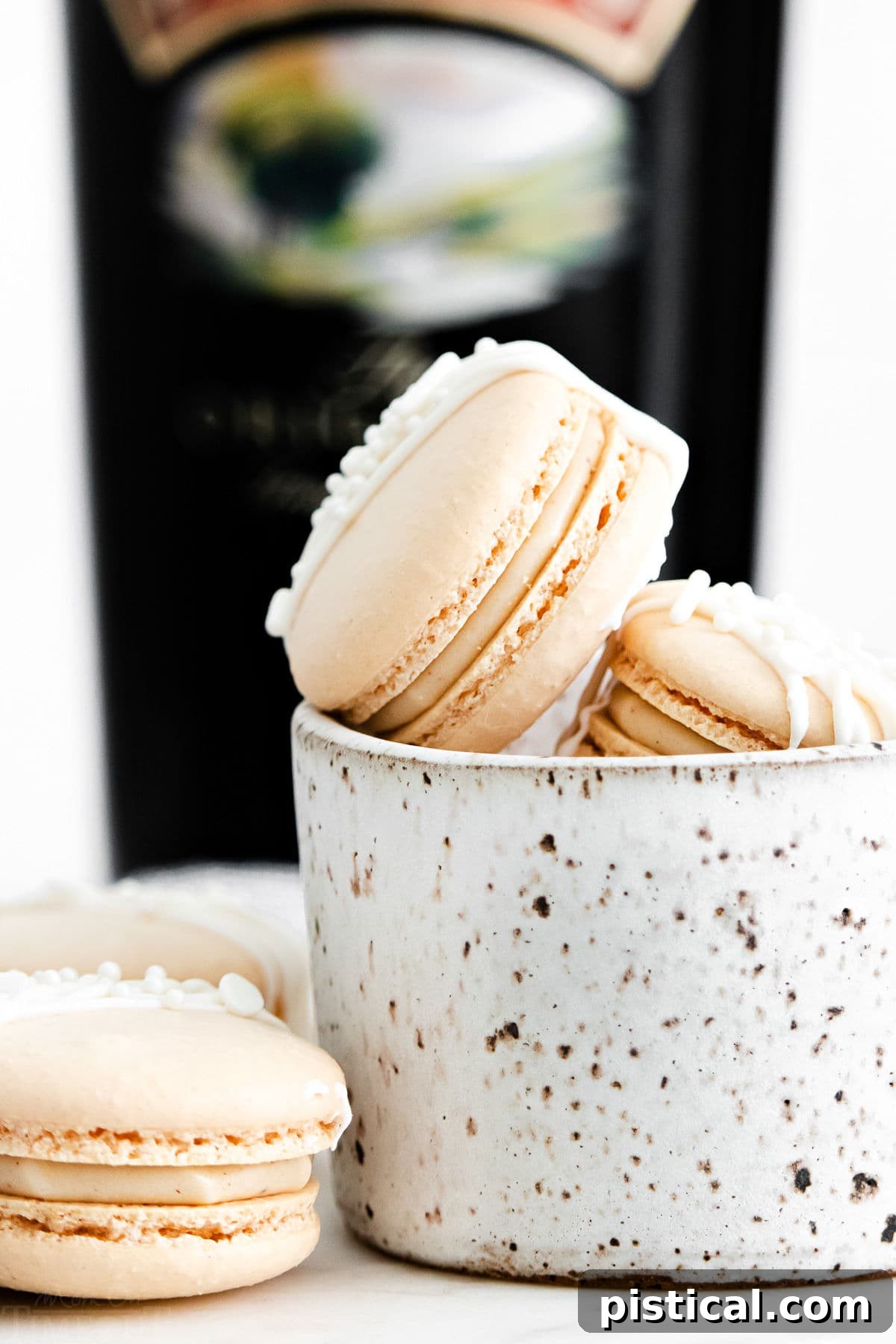 Baileys Macarons 2 Close-up of delicate Irish Cream Macarons with a rich, creamy filling, arranged next to a small white bowl and a bottle of Bailey's Irish Cream in the background. The macarons feature a perfect 'foot' and a smooth, appealing shell.