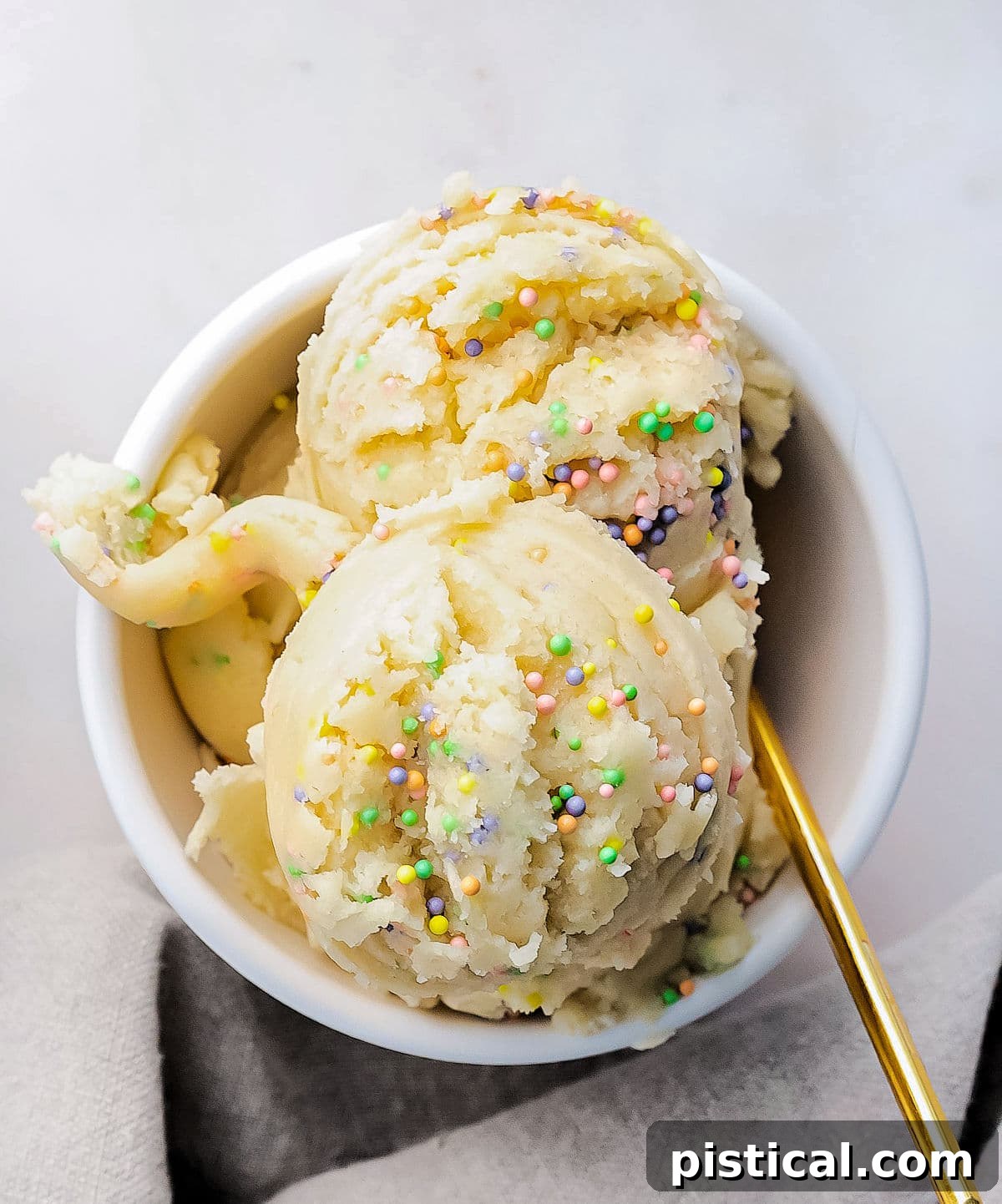 Ready-to-Eat Sugar Cookie Dough 3 Top-down view of edible sugar cookie dough in a small white bowl, adorned with colorful sprinkles and a gold spoon resting beside it.