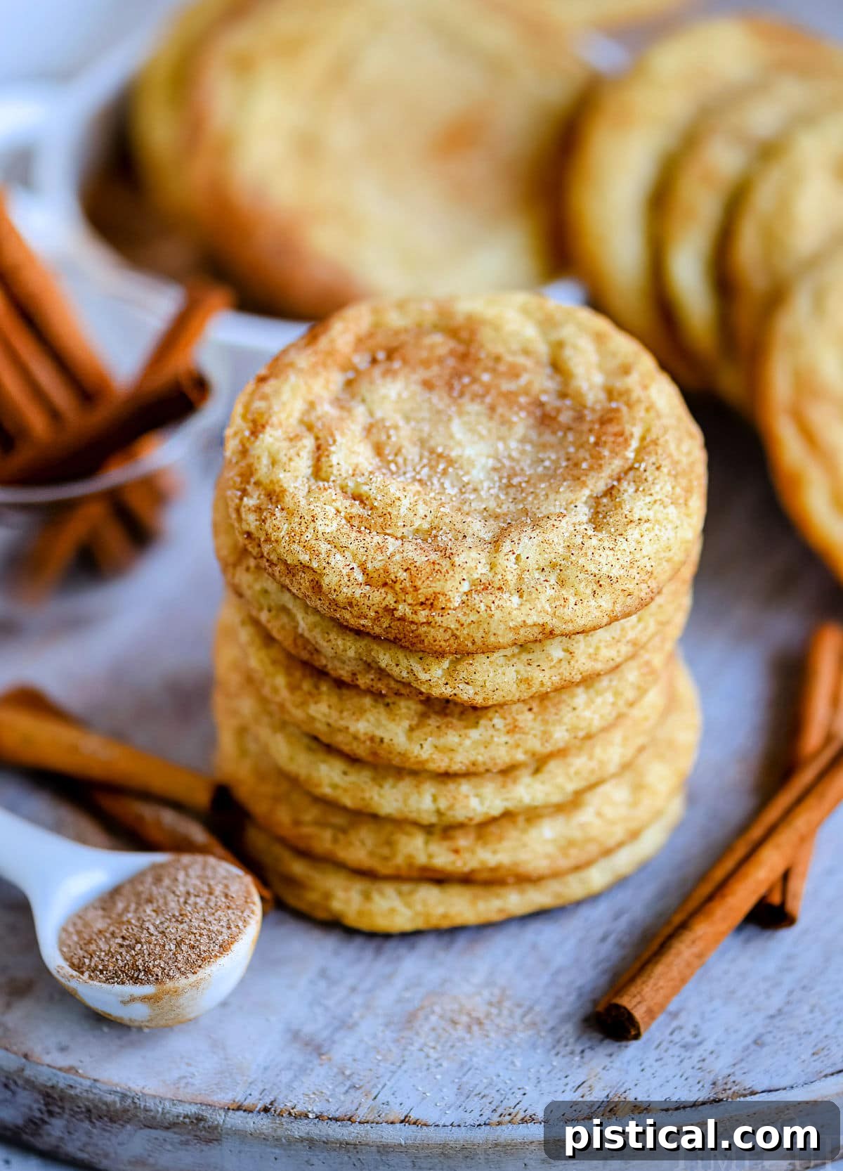 Six perfectly baked snickerdoodle cookies stacked on a blue board with cinnamon sticks scattered nearby.