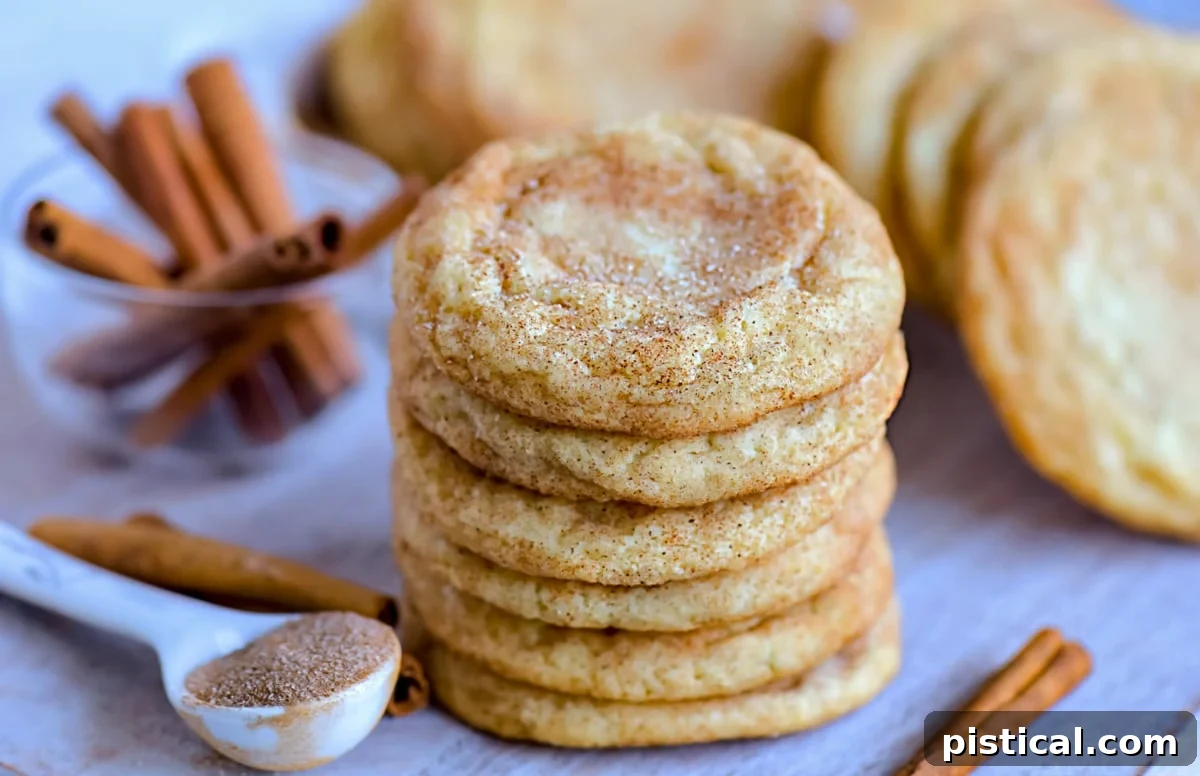Snickerdoodle cookies stacked on a wooden cutting board with more cookies in the background and cinnamon sticks scattered around.