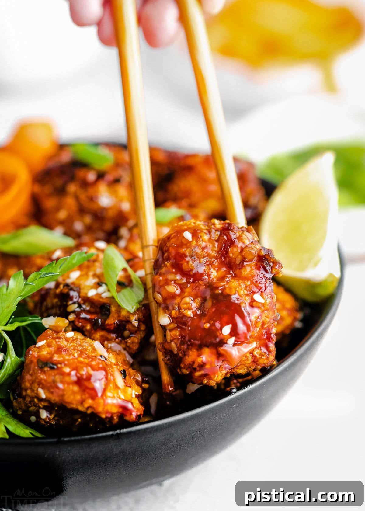 A piece of crispy teriyaki chicken being lifted from a bowl with chopsticks, showcasing its glossy sauce coating.