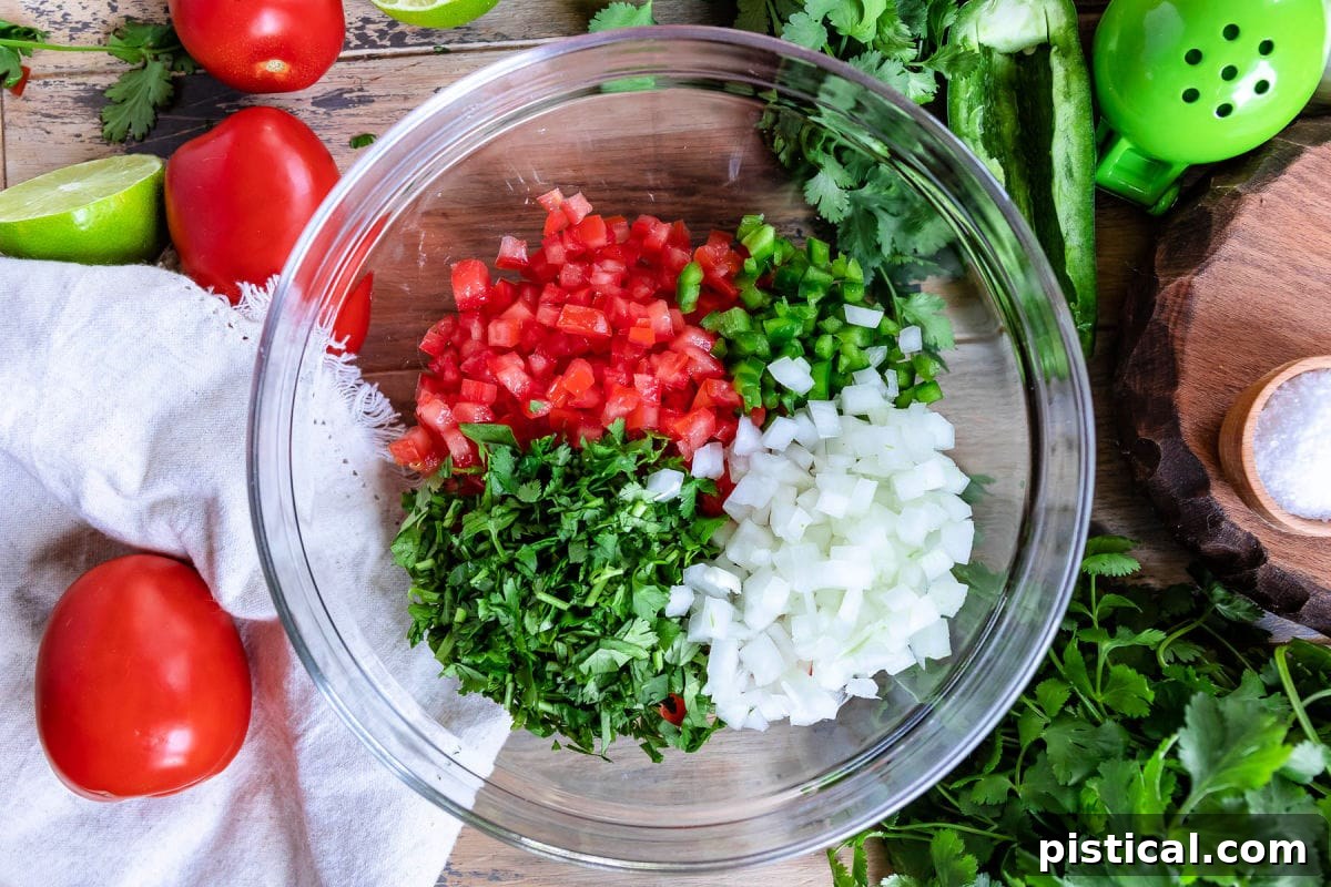 Chopped tomatoes, cilantro, jalapeño, and onion are gathered in a clear glass bowl, showcasing the fresh, raw ingredients just before mixing for Pico de Gallo.