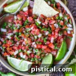 Top-down view of Pico de Gallo in a clay bowl garnished with lime wedges and surrounded by chips.