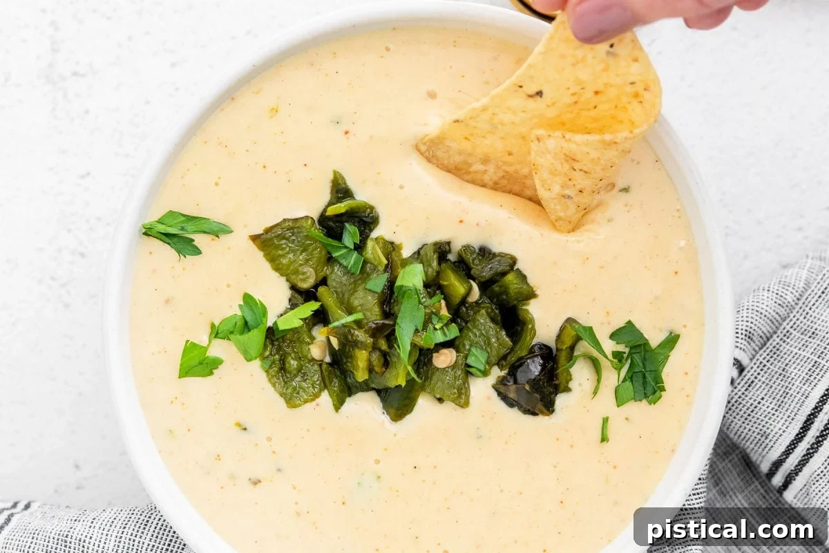 Overhead view of a white bowl filled with creamy Queso Blanco, garnished with fresh cilantro and finely diced green peppers. A hand holds a tortilla chip dipping into the rich cheese sauce.